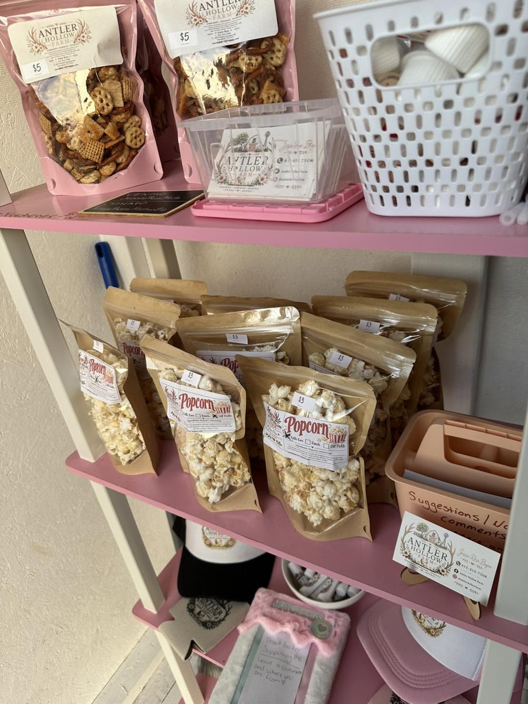 Bags of farm stand popcorn on a pink shelf beneath snack mixes.